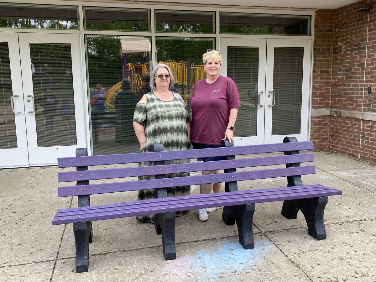 Two women standing next to purple recycled bench