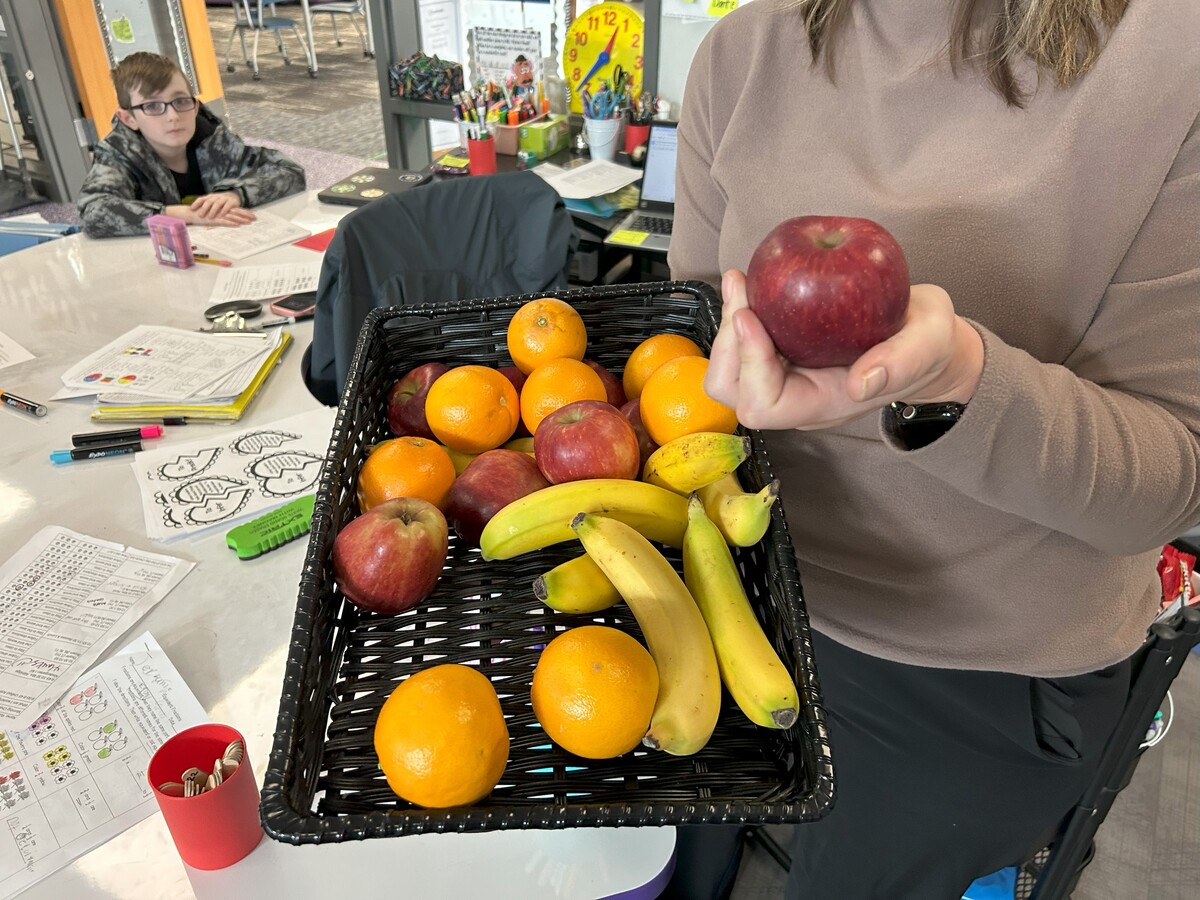 Teacher holds basket of fresh fruit in one hand, and an apple in the other.