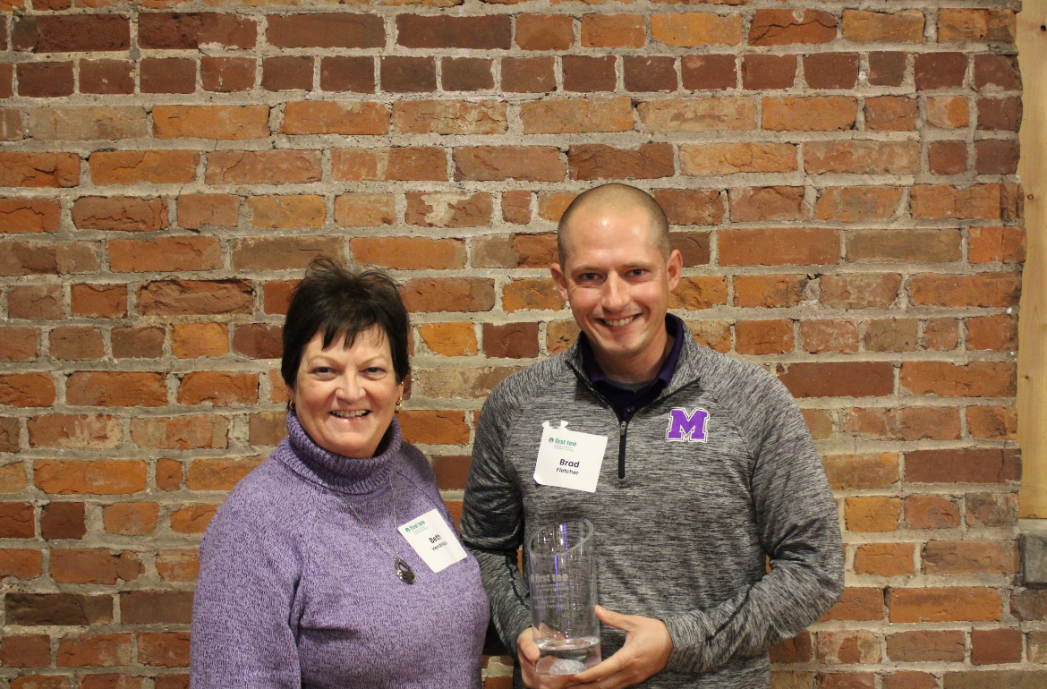 Two people smile for photo. Man, on the right, holds up award he's just won.