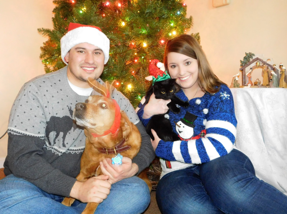 man and woman holding dog and cat in front of Christmas tree